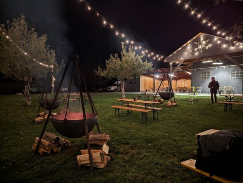 A cozy outdoor space at night with string lights, fire pits, and wooden benches, creating a warm atmosphere.
