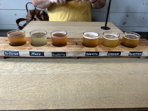 A wooden flight board with six small glasses of different colored beverages, labeled with their names.