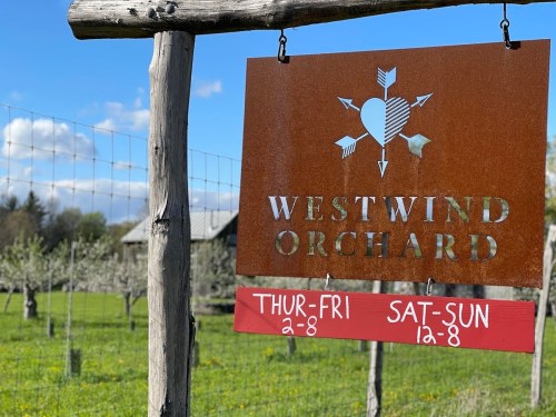 Sign for Westwind Orchard with operating hours: Thu-Fri 2-8, Sat-Sun 12-8, surrounded by trees and a blue sky.