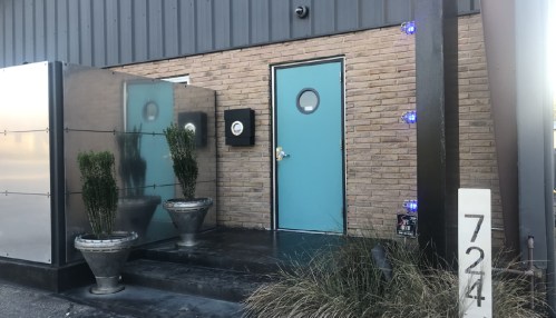 A modern entrance with a turquoise door, potted plants, and a sleek facade, featuring a brick wall and metal accents.