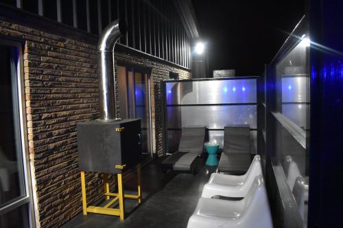 A dimly lit outdoor space featuring a black stove, two lounge chairs, and a green stool against a brick wall.