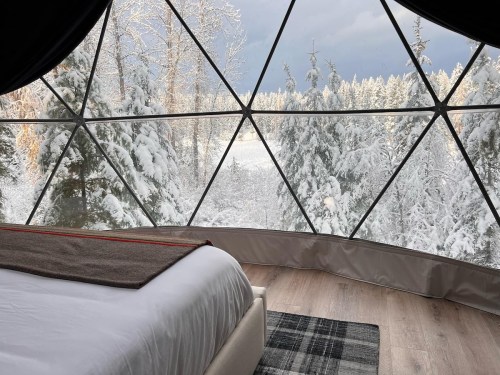Cozy interior of a geodesic dome with a bed, overlooking a snowy forest landscape through large windows.