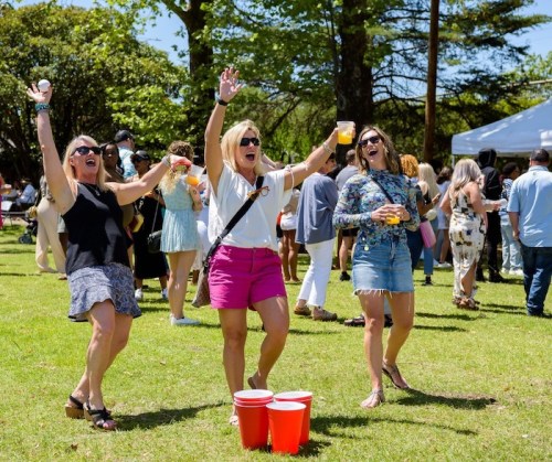 Three women joyfully celebrate outdoors, raising their arms and drinks, with a crowd and trees in the background.
