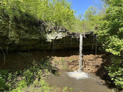 A small waterfall cascades down a rocky cliff surrounded by lush green foliage and trees.