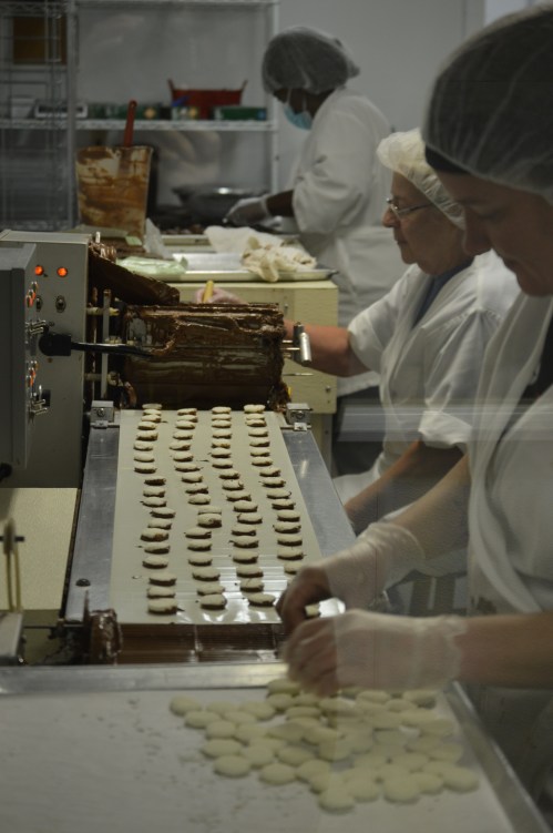 Workers in a bakery use machinery to produce cookies, with dough and chocolate filling visible on the conveyor belt.