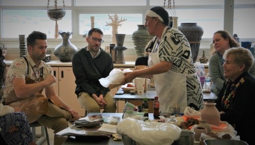A group of people watches as a woman demonstrates pottery techniques in a classroom setting.