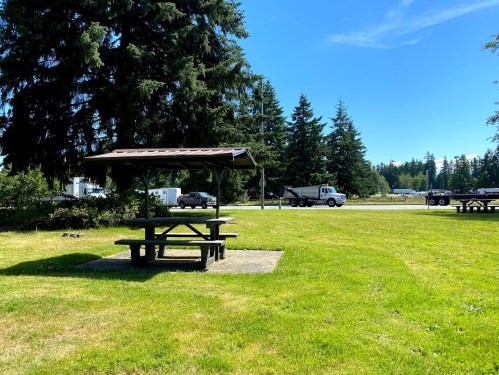A picnic area with a table and shelter, surrounded by grass and trees, near a parking area and a road.