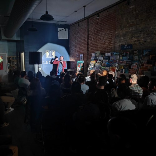 A performer on stage in a bookstore, with an audience seated in front, illuminated by a spotlight.
