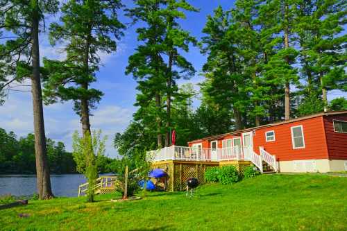 A cozy red cabin by a lake, surrounded by tall trees and green grass, with a deck and outdoor seating.