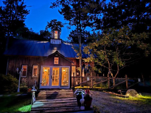 A cozy wooden cabin illuminated at dusk, surrounded by trees and a gravel path leading to its welcoming entrance.