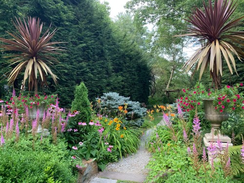 A lush garden path lined with vibrant flowers and tall plants, leading through greenery and decorative urns.