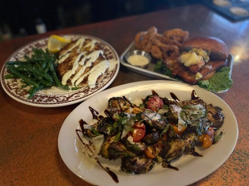A plate of roasted Brussels sprouts with tomatoes, a grilled fish dish with green beans, and a fried chicken sandwich.