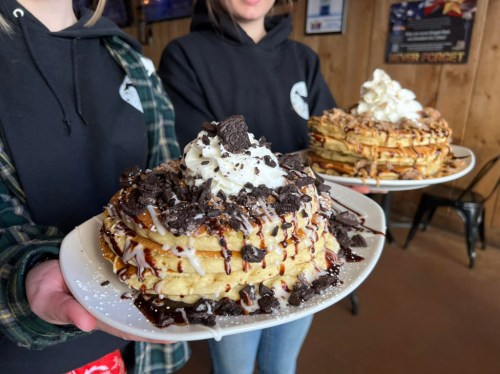 Two people hold large, stacked waffle desserts topped with whipped cream, chocolate drizzle, and crushed cookies.