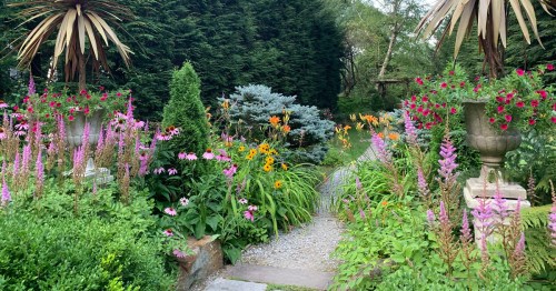 A vibrant garden path lined with colorful flowers, greenery, and decorative planters under a sunny sky.