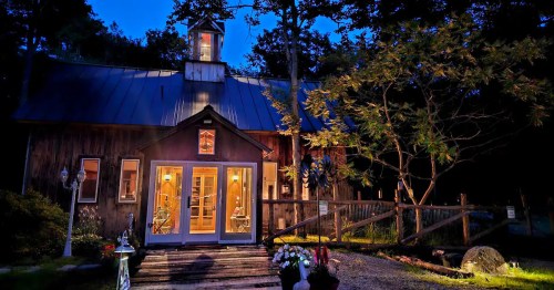 A cozy wooden cabin at dusk, with warm lights glowing from the windows and surrounded by trees and flowers.