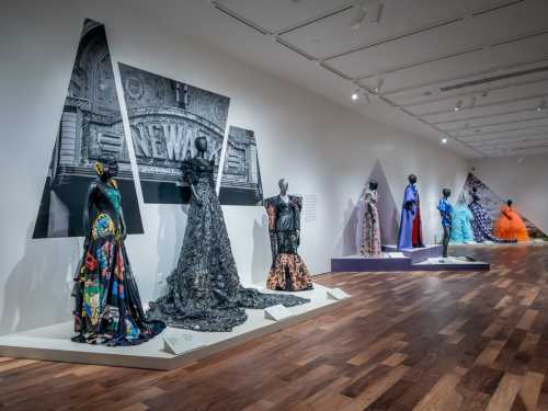 A gallery display featuring elegant dresses on mannequins, with a large black-and-white backdrop and wooden flooring.