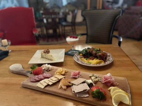 A wooden platter with assorted meats and cheeses, a dessert, and a main dish with greens and meat, all elegantly presented.