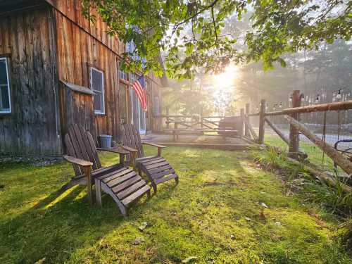 Two wooden chairs sit on a grassy area beside a rustic barn, with sunlight filtering through trees and an American flag nearby.