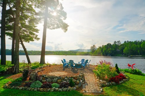 A serene lakeside scene with blue chairs, a stone path, and lush greenery under a bright sky.
