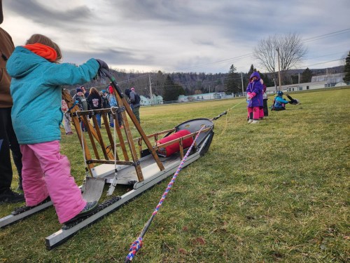 Children participate in a sledding activity on a grassy field, with a sled being pulled by one child.