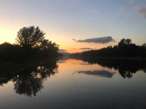 A serene river at sunset, reflecting trees and clouds in calm waters under a colorful sky.