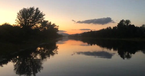 A serene sunset over a calm river, with trees and mountains reflected in the water.