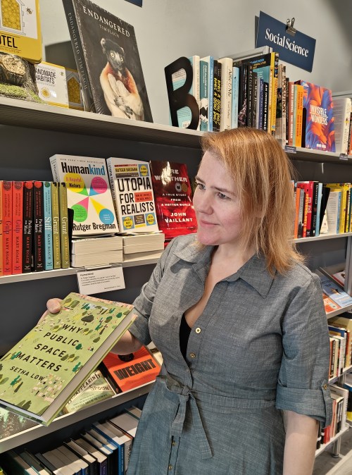 A woman holds a book titled "Public Space Matters" in a bookstore, surrounded by shelves of various books.