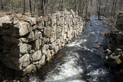 A stone wall beside a flowing stream, surrounded by trees and rocky terrain in a natural setting.