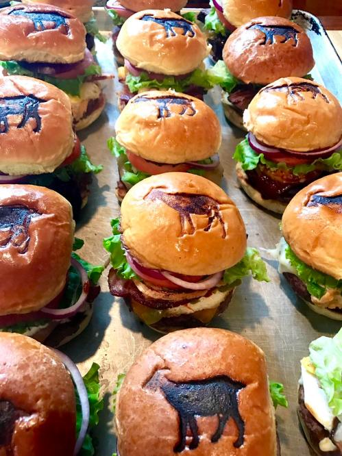 A tray of burgers with lettuce, tomato, and onion, featuring a goat logo stamped on the buns.