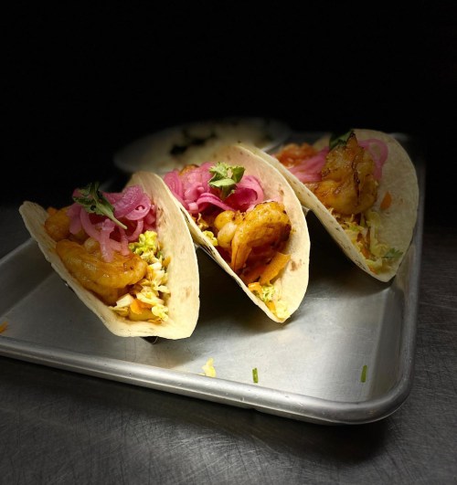 Three shrimp tacos on a metal tray, topped with pink pickled onions and garnished with cilantro.
