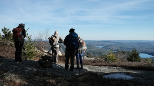 A group of hikers stands on a rocky outcrop, gazing at a scenic view of mountains and lakes under a clear blue sky.