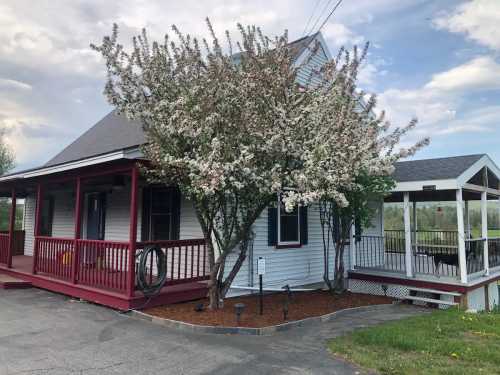 A white house with a red porch and a flowering tree in front, alongside a gazebo in a grassy area.