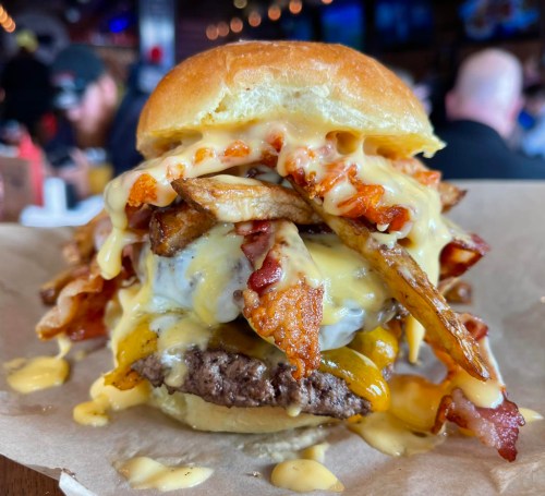 A towering burger with melted cheese, crispy bacon, and fries stacked on top, served on a soft bun.