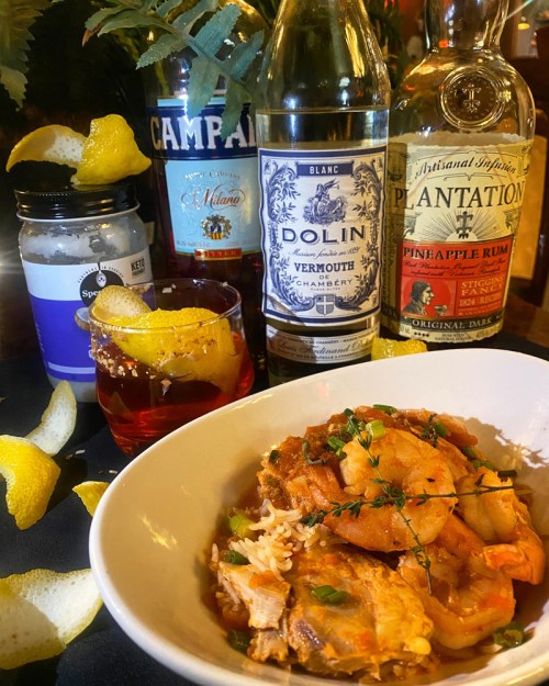 A bowl of shrimp and rice dish with citrus peels and various bottles of spirits in the background.