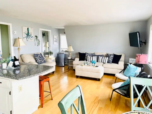 Bright living room with a beige sofa, decorative pillows, a TV, and a dining area featuring a green chair and a round table.