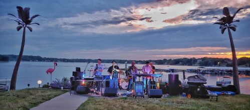 A band performs by a lake at sunset, with palm trees and a flamingo decoration nearby.