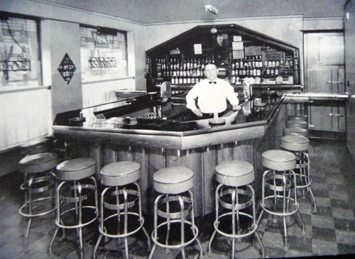 A vintage black-and-white photo of a bar with a bartender standing behind the counter and stools arranged in front.