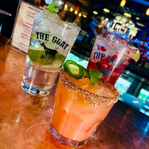 Three colorful cocktails on a bar counter, featuring a salted rim drink, a clear drink, and a red drink in branded glasses.