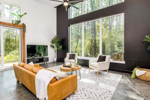 Modern living room with large windows, a leather sofa, two chairs, and a view of a forested area outside.