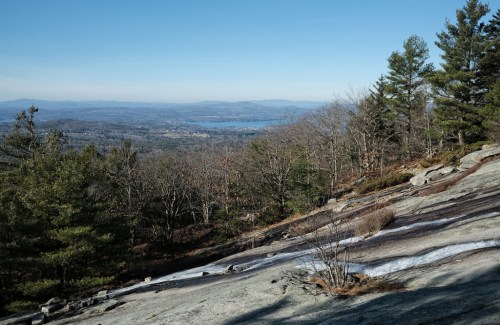 A scenic view from a rocky hillside, showcasing a lake and distant mountains under a clear blue sky.