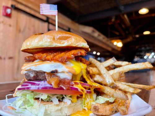 A towering burger with cheese, bacon, and onion, topped with a flag, served with crispy fries on the side.