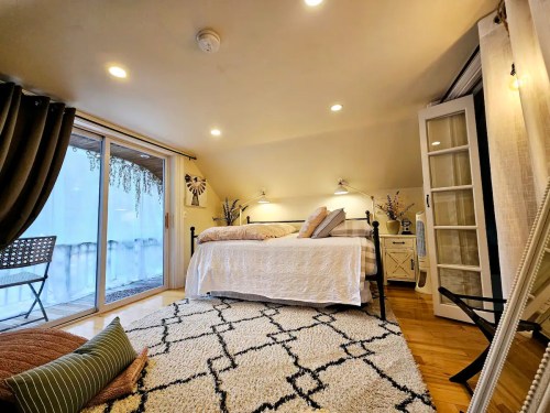Cozy bedroom with a large bed, soft lighting, and a sliding glass door leading to a balcony. Decorated with plants and rugs.