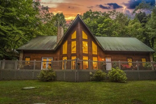 A cozy log cabin surrounded by trees, with large windows glowing at sunset.