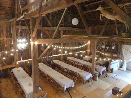 A rustic barn interior with wooden beams, string lights, and long tables set for an event.