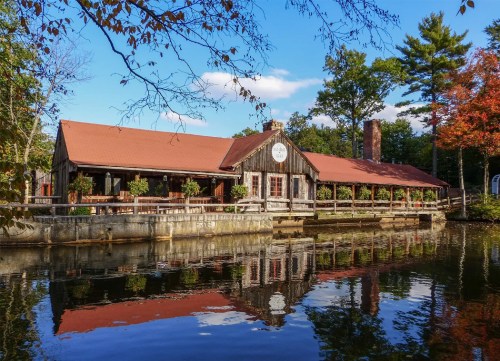 A rustic wooden building with a red roof reflects in calm water, surrounded by trees and autumn foliage.