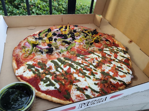 A large pizza in a box, half topped with olives, peppers, and onions, the other half with pesto and fresh mozzarella.