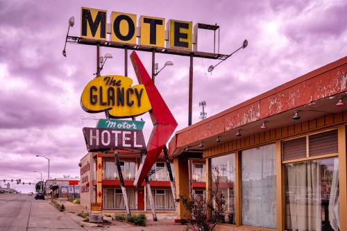 A vintage motel sign with "The Glancy Motor Hotel" under a cloudy sky, showcasing retro architecture and faded colors.