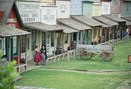 A historic street with colorful wooden storefronts, a horse-drawn wagon, and two people in period costumes.