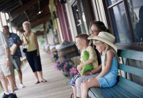 Two children sit on a bench, one holding a green toy, while an adult smiles beside them. People walk in the background.