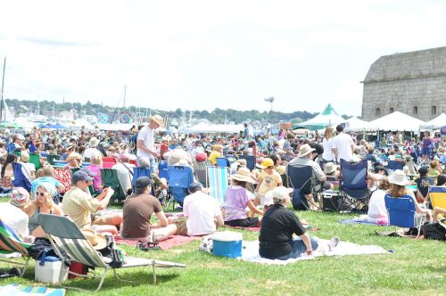 A large crowd of people sitting on the grass at an outdoor event, with tents and boats in the background.